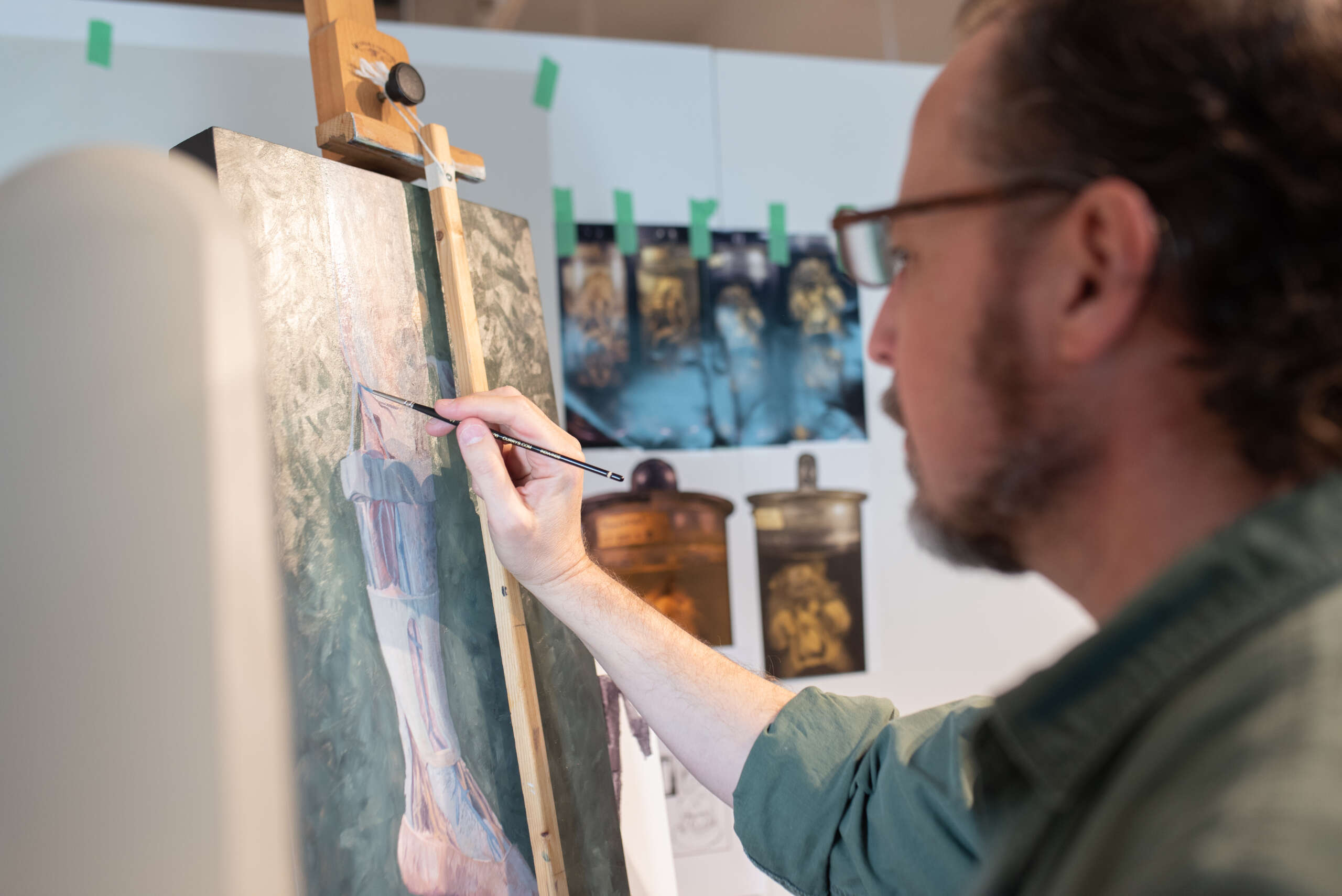 Man painting on an easel with paintings hanging on the wall.